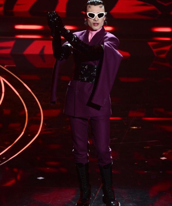 <p>Eleganza <strong>fetish</strong> per l&rsquo;artista pi&ugrave; chiacchieratə del Festival numero 73. Sul palco si esibisce in un <strong>doppiopetto bordeaux</strong> con maniche penzolanti, indossato sopra un <strong>dolcevita in latex rosa</strong> e strizzato in vita da una cintura corsetto. Gli accessori non sono da meno: occhiali da sole sagomati, lunghi guanti di vernice nera e combat boots stringati.</p>
